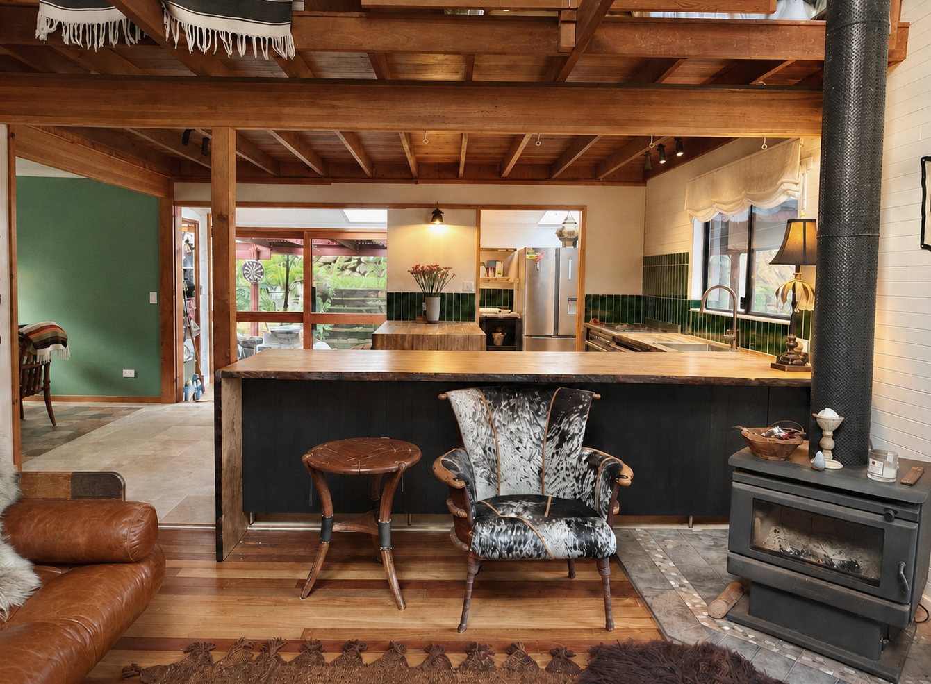The kitchen through the living room: a long timber bench, green tiled splashback, exposed beams and a wood heater